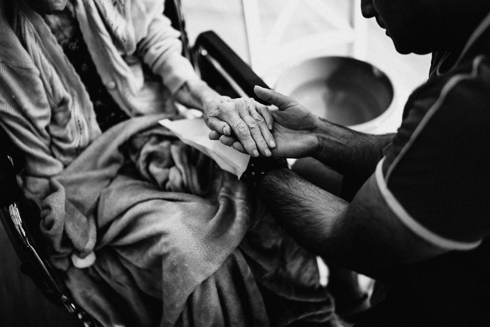 A caregiver holds the hand of an elderly woman seated under a blanket, in black and white.
