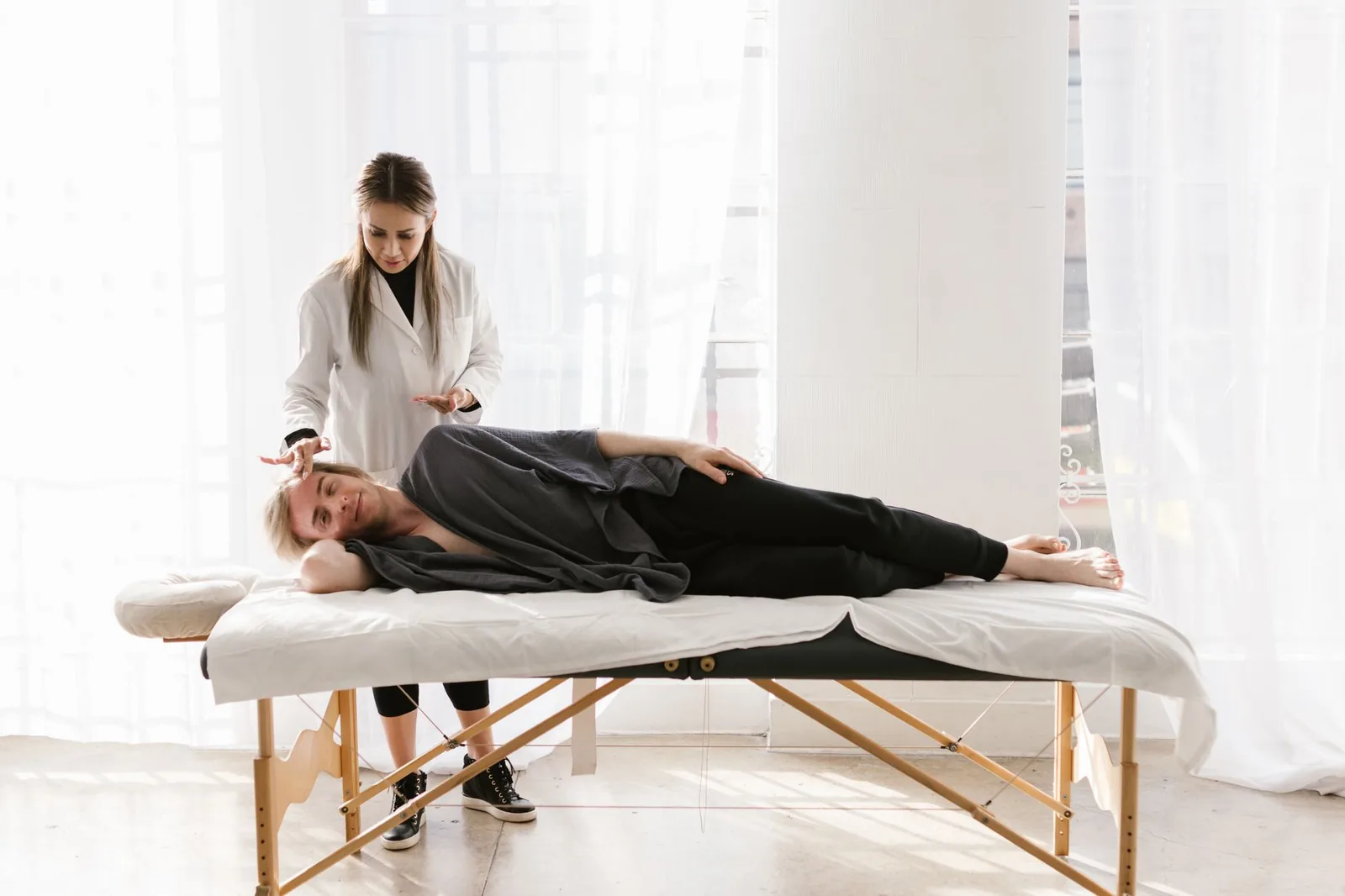 A practitioner working with her hands held above a fully clothed client resting on a portable table in a bright room.