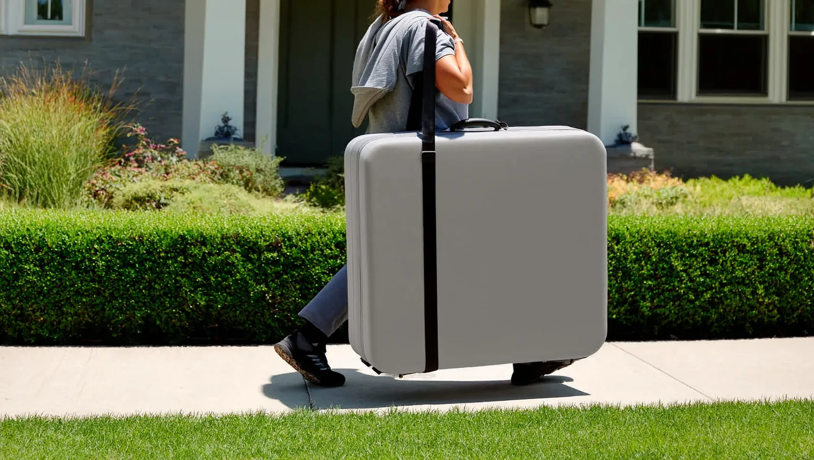 A mobile massage therapist carrying a folded portable massage table in a padded case toward a client's front door.